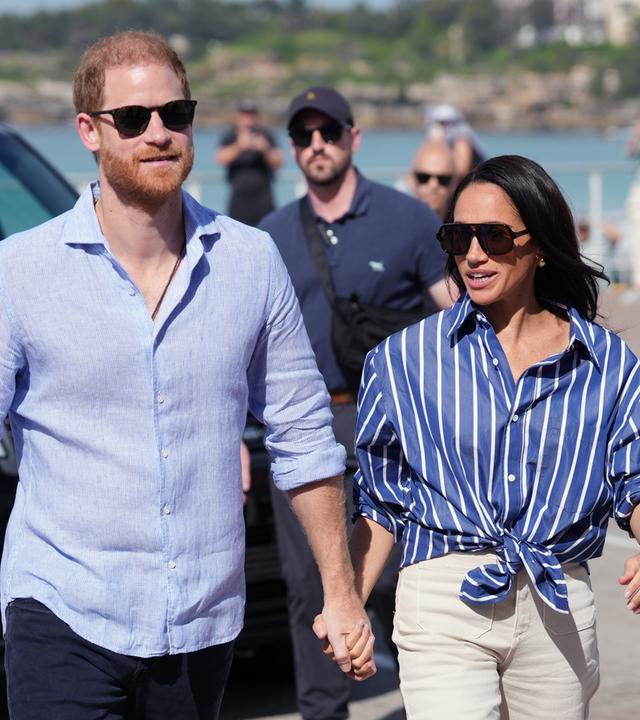 Auf dem Bild zu sehen ist: Meghan und Harry Hand in Hand eine Straße entlang laufen. Die Perspektive ist eine Halbtotale.