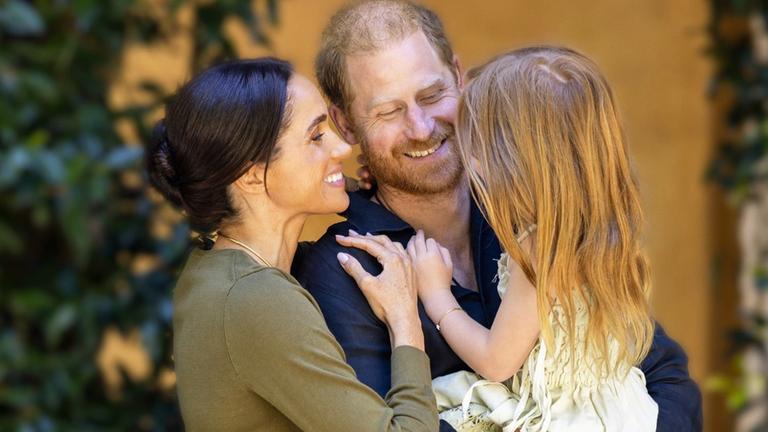 Prinz Harry hält ein Kind mit langen blonden Haaren auf den Armen, Meghan Markle lächelt es an.