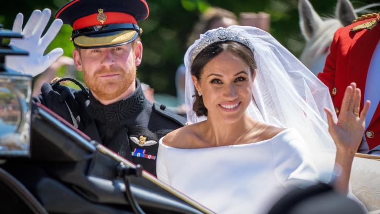 Meghan Markle und Prinz Harry winken in Hochzeitskleidung aus einer Kutsche.