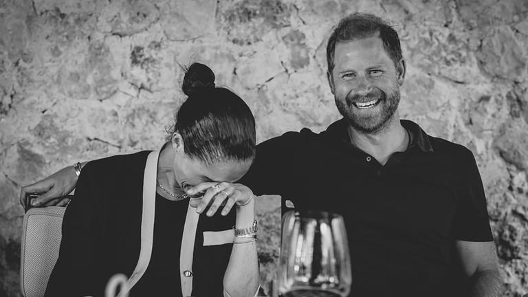 Meghan (links) und Harry sitzen an einem Tisch und lachen. 