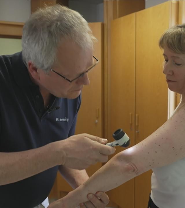 Ein Arzt untersucht mit einer Lupe eine Patientin auf Hautkrebs.