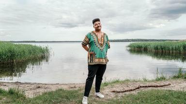 Dennis, ein junger Mann mit dunklem Teint, steht am Ufer eines ruhigen Sees, der Himmel ist größtenteils bewölkt. Er steht auf einer kleinen Grasfläche, die Hände sind locker in die Hosentaschen gesteckt, die Körperhaltung wirkt entspannt und freundlich.