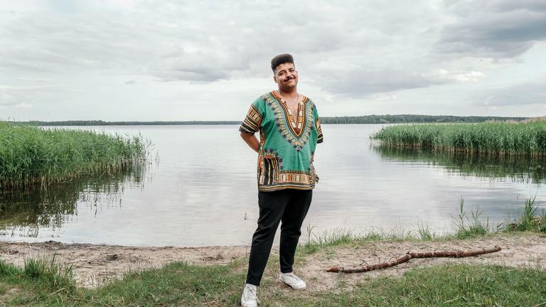 Dennis, ein junger Mann mit dunklem Teint, steht am Ufer eines ruhigen Sees, der Himmel ist größtenteils bewölkt. Er steht auf einer kleinen Grasfläche, die Hände sind locker in die Hosentaschen gesteckt, die Körperhaltung wirkt entspannt und freundlich.
