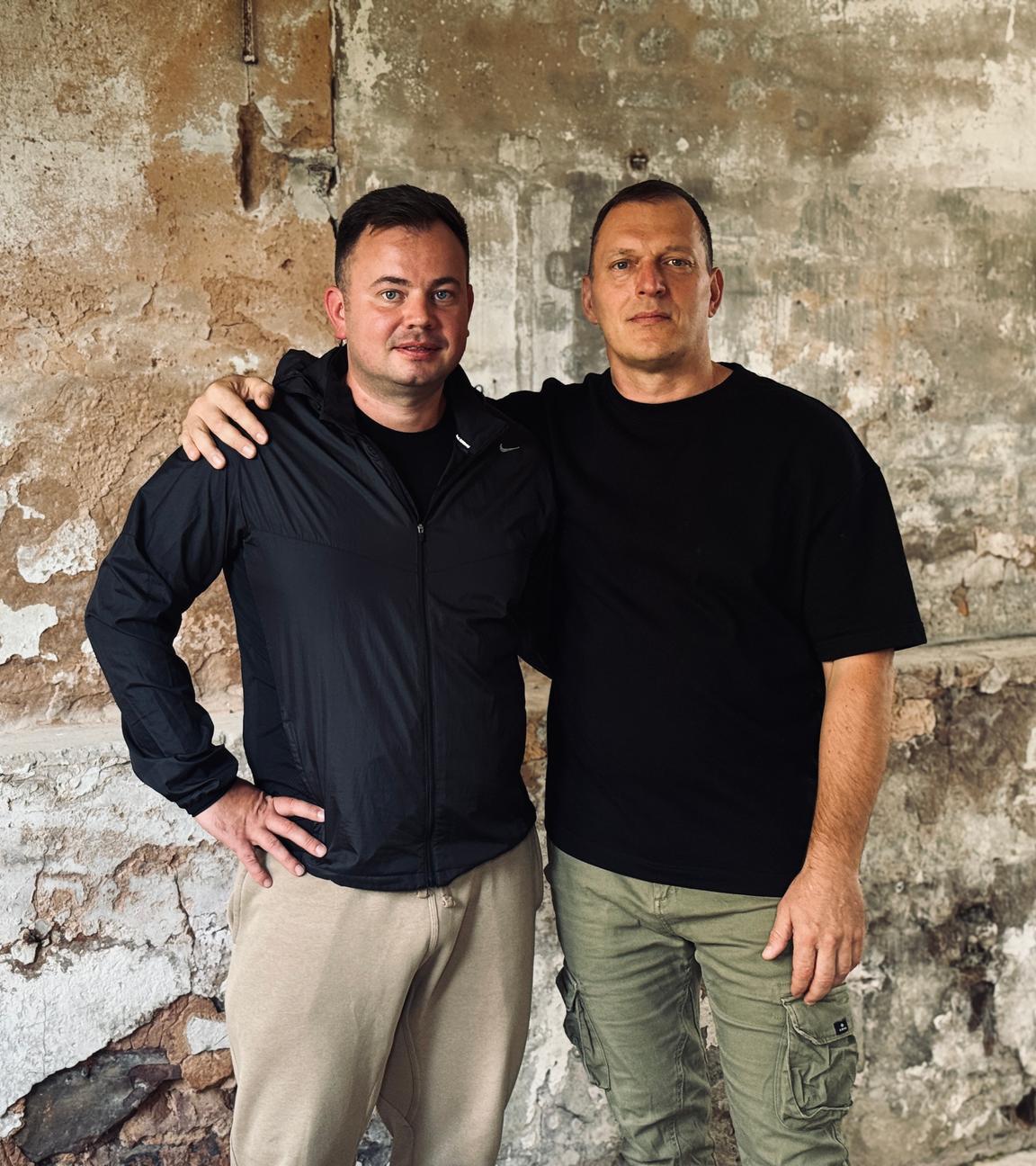 Vitali Litz und Sergej Mermis stehen eng nebeneinander vor einer alten Steinwand. Sergej hat Vitali seine Hand auf die Schulter gelegt.