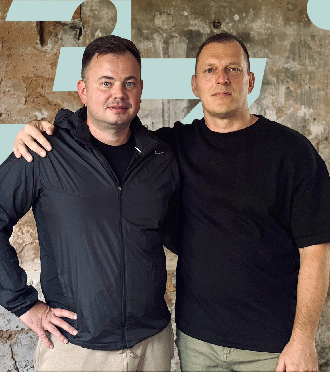 Vitali Litz und Sergej Mermis stehen eng nebeneinander vor einer alten Steinwand. Sergej hat Vitali seine Hand auf die Schulter gelegt.