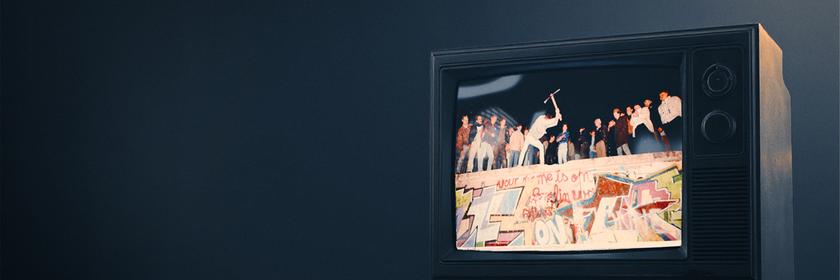 Ein alter Röhrenfernseher, der den Sturz der mauer zeigt. Auf ihr stehen mehrere Personen und feiern die Wiedervereinigung. 