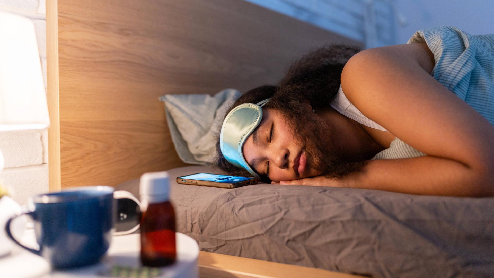 Eine junge Frau mit Schlafmaske liegt schlafend im Bett. Vor ihr auf einem Nachttisch befindet sich eine Flasche, eine Tasse und Tabletten.