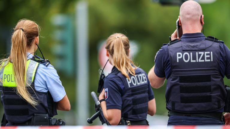 Polizeibeamte stehen zur Absicherung an einer Absperrung am Tatort. Nach Schüssen am 22.08.2025 in Menden im Sauerland ist die Polizei im Großeinsatz.