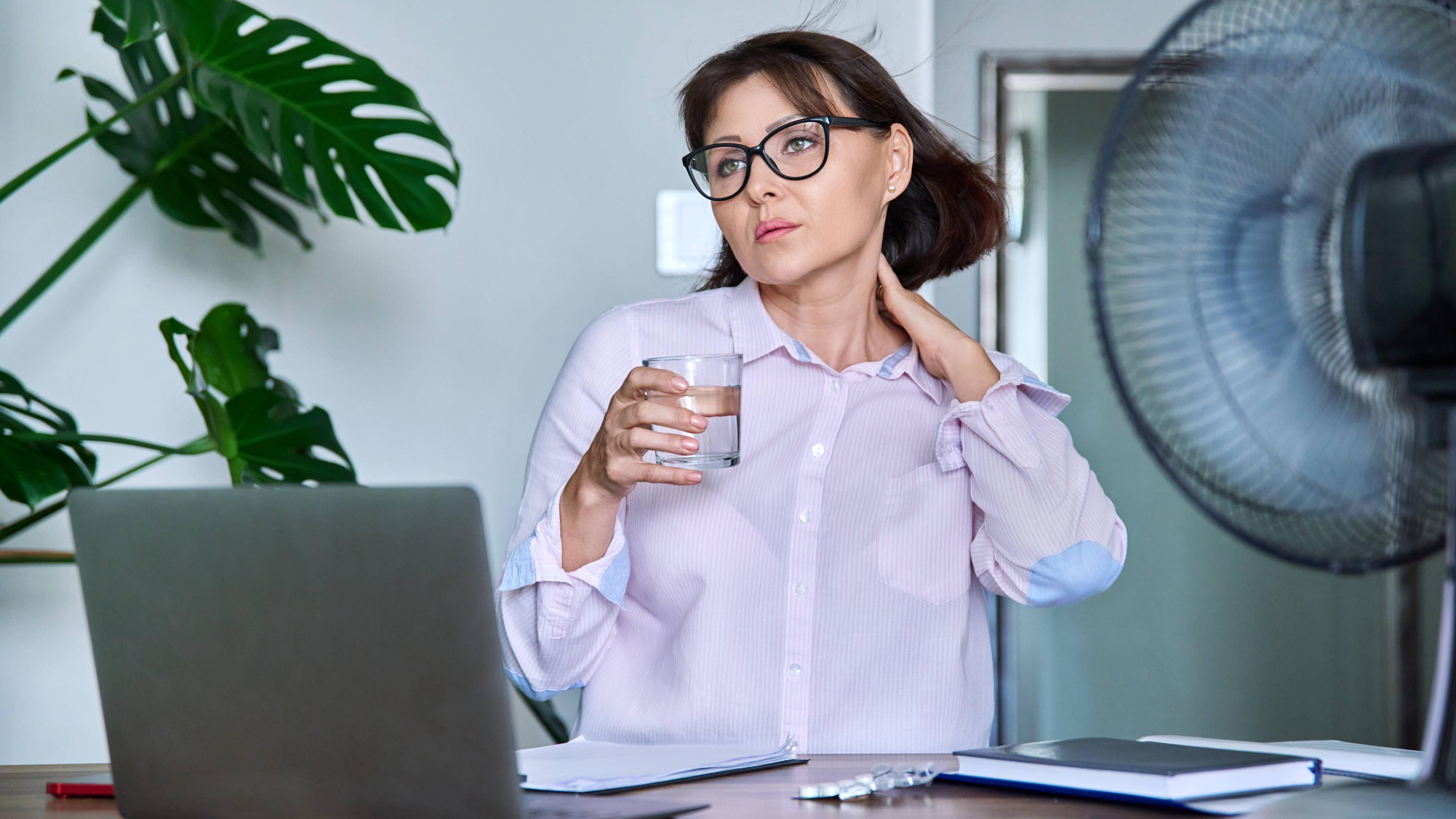 Eine Frau am Schreibtisch hält ein Glas Wasser in der Hand und fasst sich an den Hals.