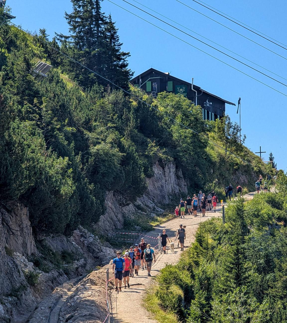 Viele Menschen bei einer Bergwanderung auf einem breiten Wanderweg Richtung Gipfel und einer Hütte.