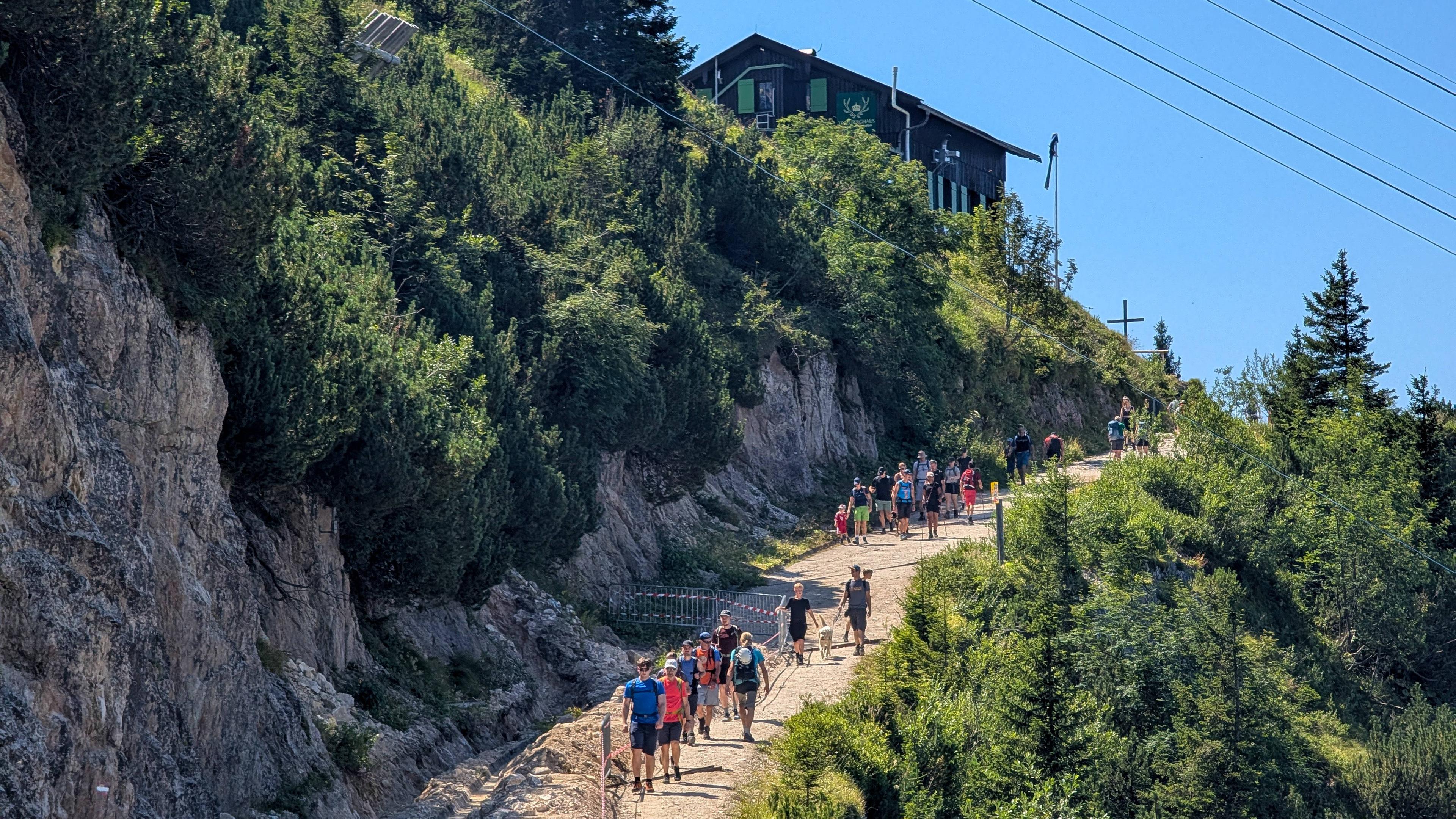 Viele Menschen bei einer Bergwanderung auf einem breiten Wanderweg Richtung Gipfel und einer Hütte.