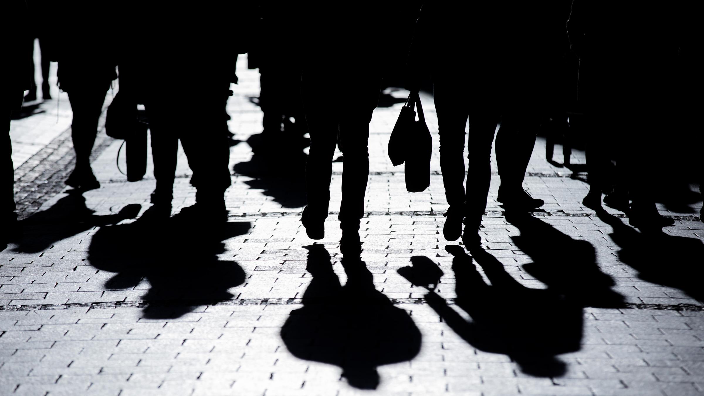 Menschen gehen in starkem Gegenlicht mit ihren Einkäufen durch die Innenstadt und werfen Schatten auf den Asphalt. 