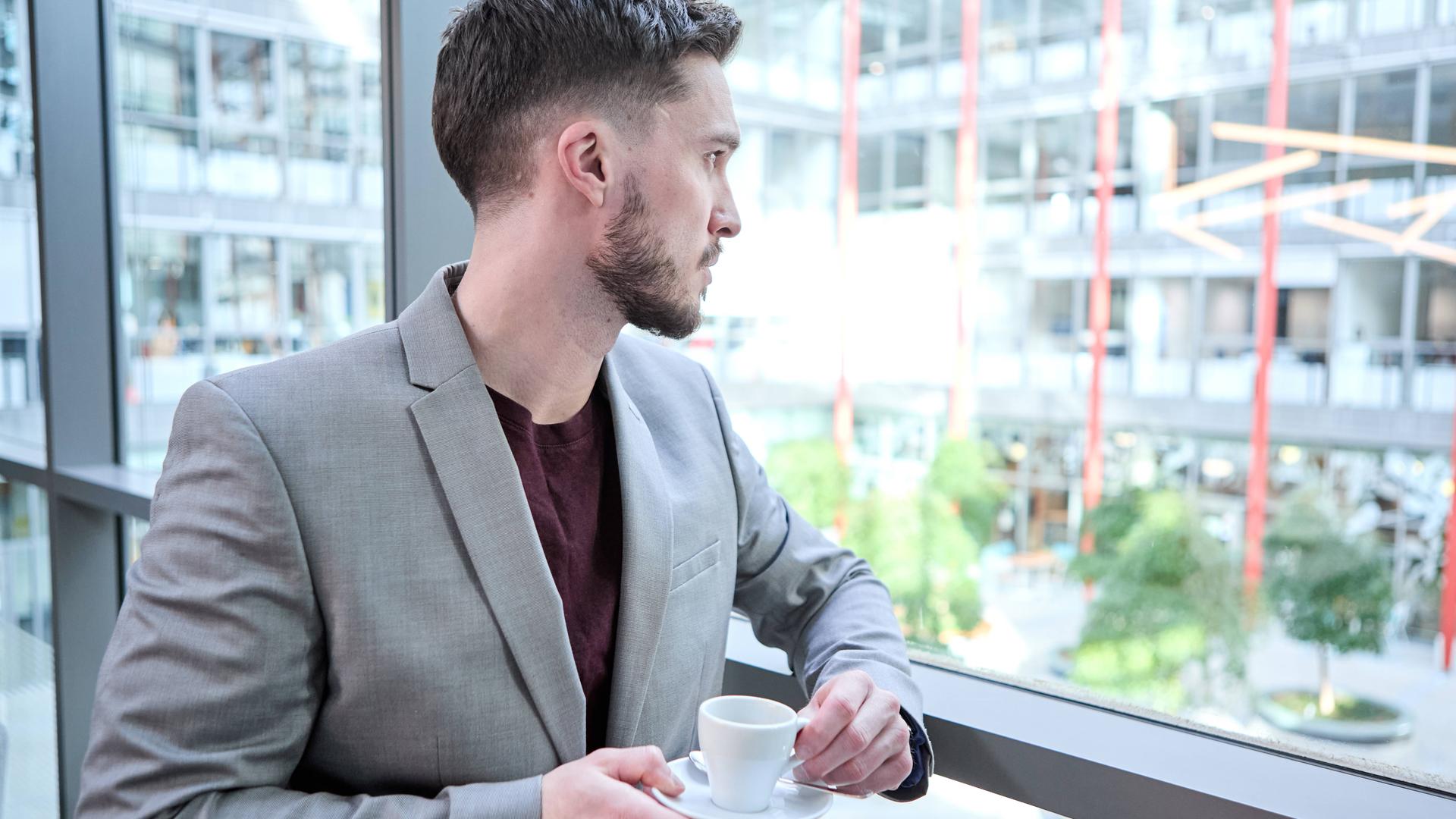 Ein junger Mann im grauen Anzug blickt nachdenklich aus einem Bürofenster, während er eine Tasse Kaffee hält.
