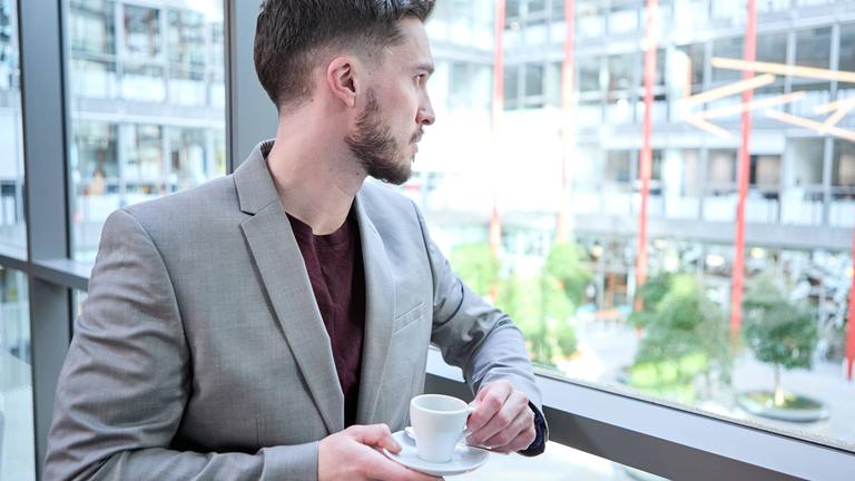 Ein junger Mann im grauen Anzug blickt nachdenklich aus einem Bürofenster, während er eine Tasse Kaffee hält.