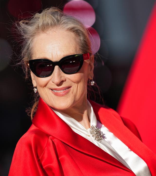 Schauspielerin Meryl Streep in roter Jacke und Sonnenbrille auf einem roten Teppich.