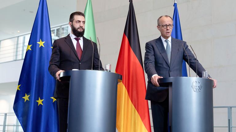 Friedrich Merz empfängt den syrischen Übergangspräsidenten: Ahmed al-Scharaa. Im Hintergrund stehen die syrische, die deutsche und die europäische Flagge