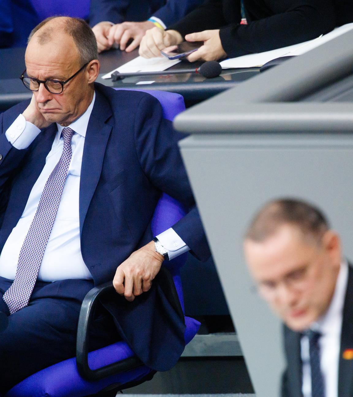 Merz im Bundestag. Im Vordergrund steht Tino Chrupalla am Podium.