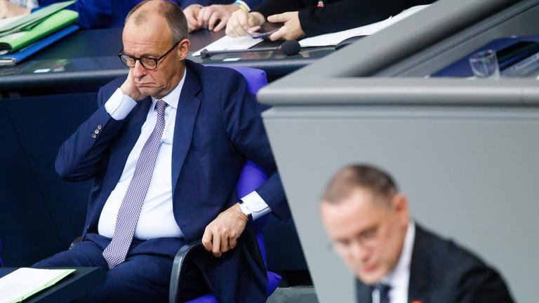 Merz im Bundestag. Im Vordergrund steht Tino Chrupalla am Podium.