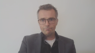 Prof. Dr. Marc Debus von der Universität Mannheim per Videocall dazugeschaltet. Er trägt eine schwarze eckige Brille und ein dunkelgraues Sakko. Hinter ihm ist nur eine weiße Wand zu sehen.