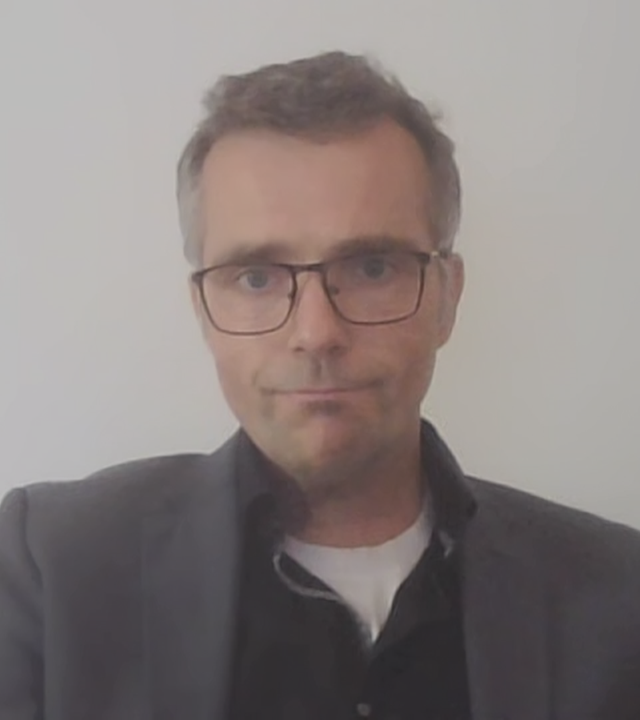 Prof. Dr. Marc Debus von der Universität Mannheim per Videocall dazugeschaltet. Er trägt eine schwarze eckige Brille und ein dunkelgraues Sakko. Hinter ihm ist nur eine weiße Wand zu sehen.