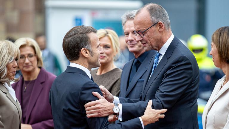  Frankreichs Präsident Emmanuel Macron (M.l) begrüßt Bundeskanzler Friedrich Merz (CDU) bei seiner Ankunft vor der saarländischen Staatskanzlei während der zentralen Feierlichkeiten dem Tag der Deutschen Einheit, Brigitte Macron, Anke Rehlinger (SPD), saarländische Ministerpräsidentin und Bundesratspräsidentin, Bundestagspräsidentin Julia Klöckner (CDU) und ihr Partner, der Fernsehmoderator Jörg Pilawa, sowie Charlotte Merz, Frau des Bundeskanzlers, sehen zu.