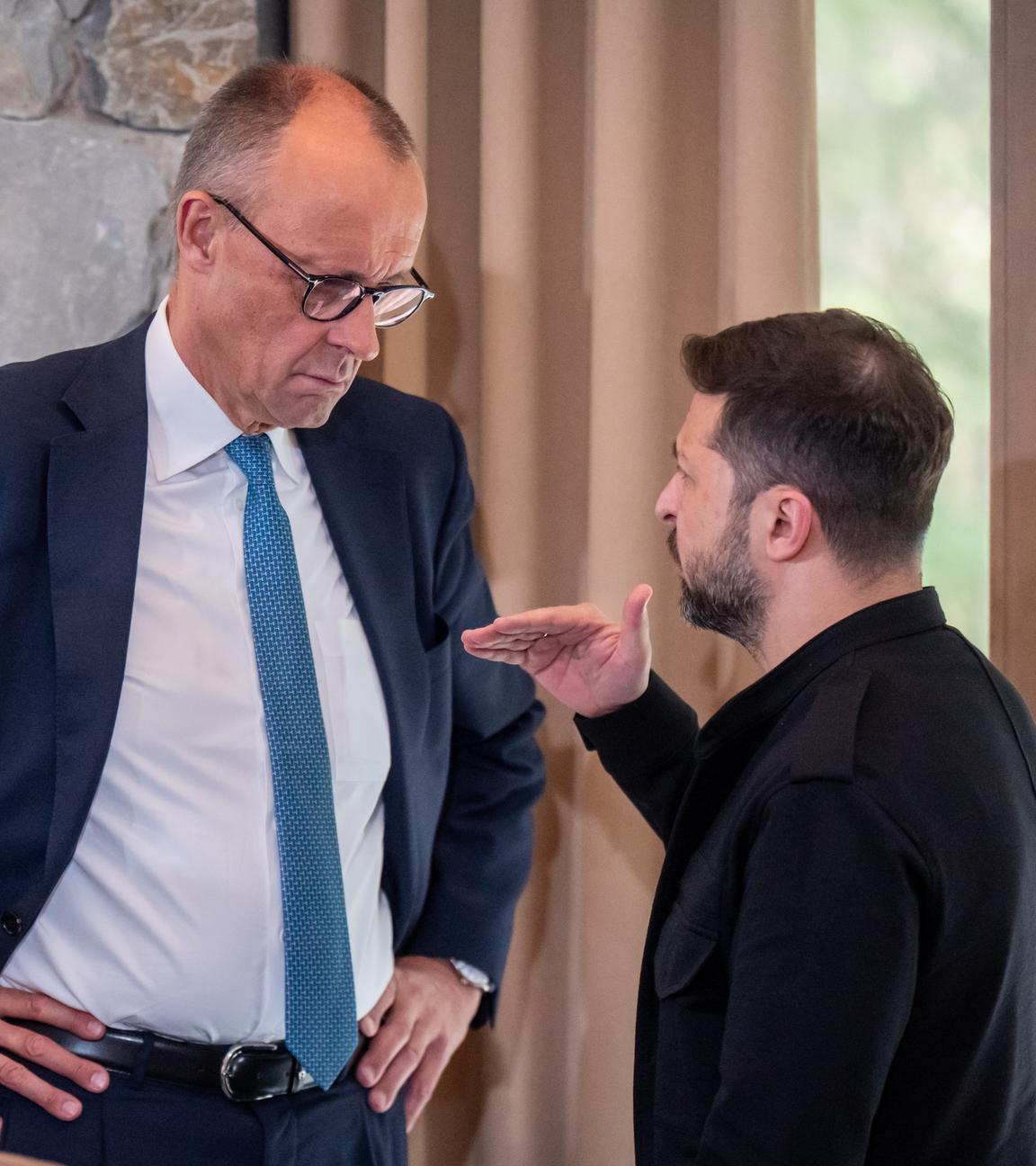 Friedrich Merz steht links im Bild, hat die Arme in die Hüfte gestemmt und hört Wolodymyr Selenskyj beim reden zu, welcher rechts im Bild steht.