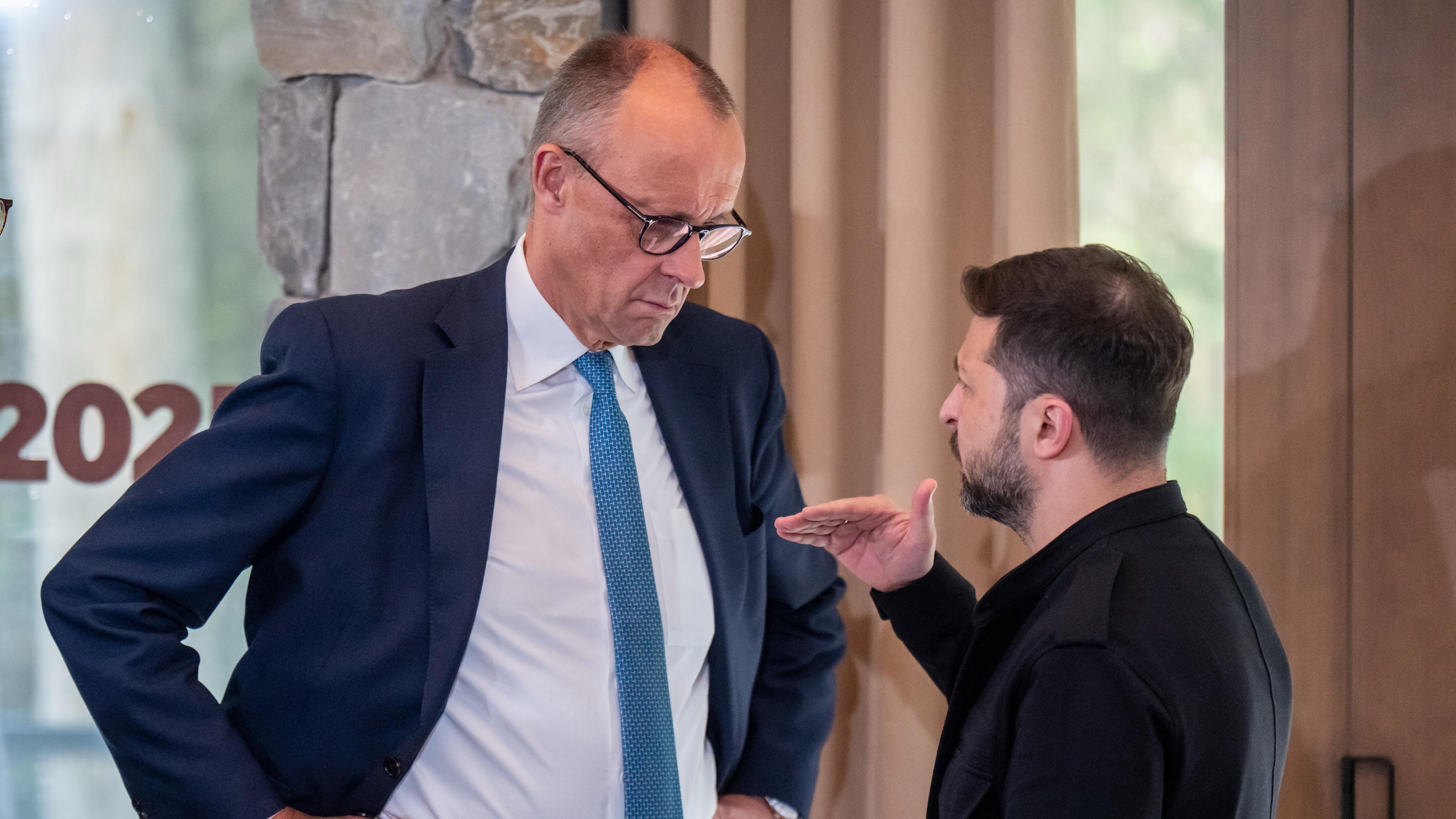 Friedrich Merz steht links im Bild, hat die Arme in die Hüfte gestemmt und hört Wolodymyr Selenskyj beim reden zu, welcher rechts im Bild steht.