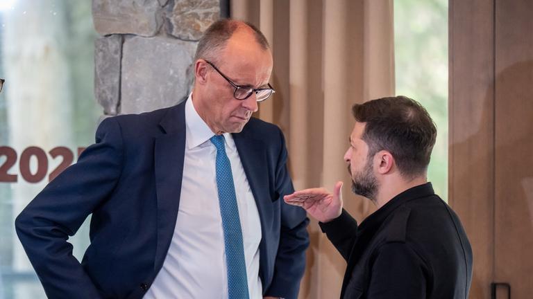 Friedrich Merz steht links im Bild, hat die Arme in die Hüfte gestemmt und hört Wolodymyr Selenskyj beim reden zu, welcher rechts im Bild steht.