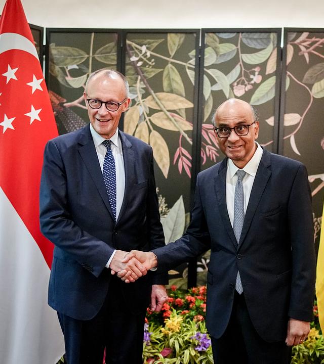 Bundeskanzler Friedrich Merz (CDU) wird vom Präsidenten der Republik Singapur, Tharman Shanmugaratnam, empfangen. Beide schauen in die Kamera und schütteln sich die Hände Im Hintergrund sieht man die Flaggen von Singapur und Deutschland.