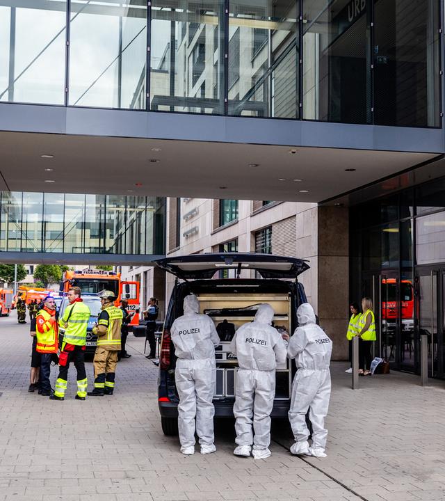 Beamte der Spurensicherung stehen vor einem Einkaufszentrum. Dahinter sind Feuerwehrmänner sowie Rettungssanitäter und Fahrzeuge zu sehen.