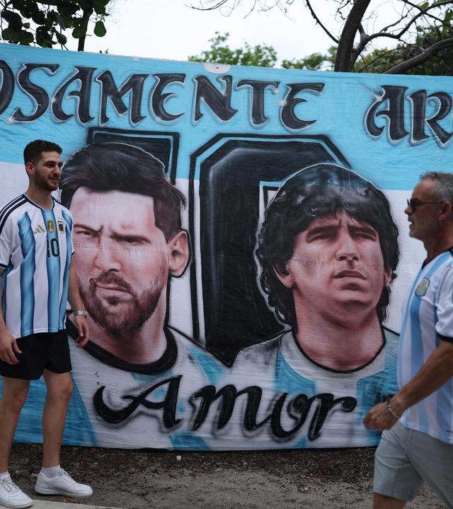 Zwei Argentinien-Fans gehen in Miami Beach an einem Plakat vorbei, auf dem die Konterfeis von Lionel Messi und Diego Maradona zu sehen ist.