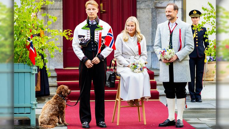 Mette Marit sitzt mit Sauerstoffgerät beim Nationalfeiertag 2026, Sverre Magnus und Haakon stehen neben ihr.