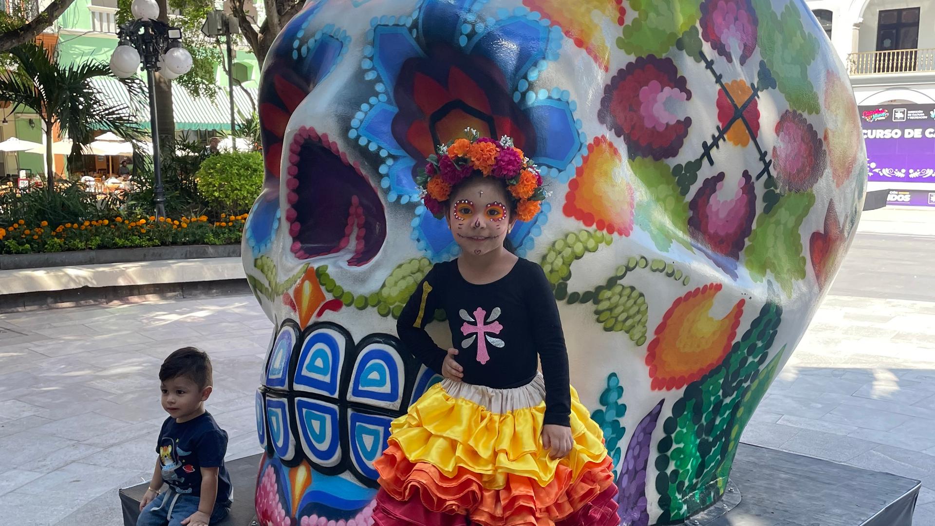 Ein Mädchen in einem bunten Kostüm und mit aufgemalter Totenmaske steht vor einem riesigen Totenkopf.