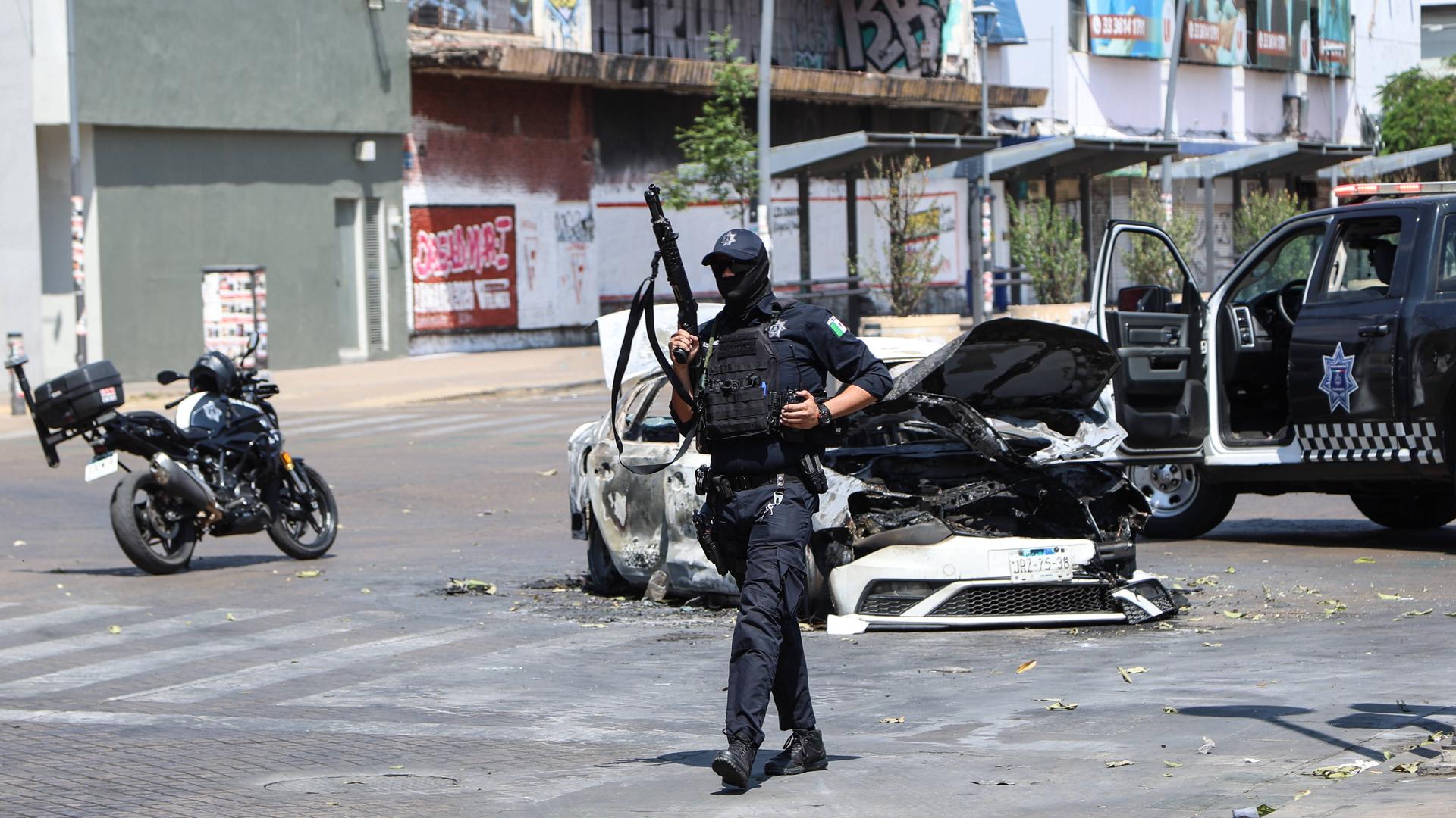 Ein Polizist bewacht am 22.02.2026 ein ausgebranntes Auto auf einer Straße in Guadalajara, der Hauptstadt des mexikanischen Bundesstaates Jalisco. 