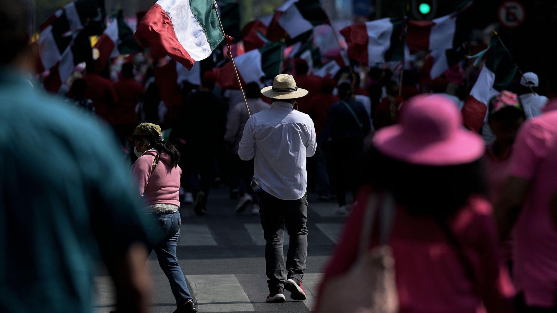 Ein Mann in einem blauen Hemd und einem Strohhut hält eine mexikanische Fahne in den Händen. Er läuft durch eine Menschenmenge. Im Hintergrund ist ein Meer an Mexiko-Flaggen zu sehen.