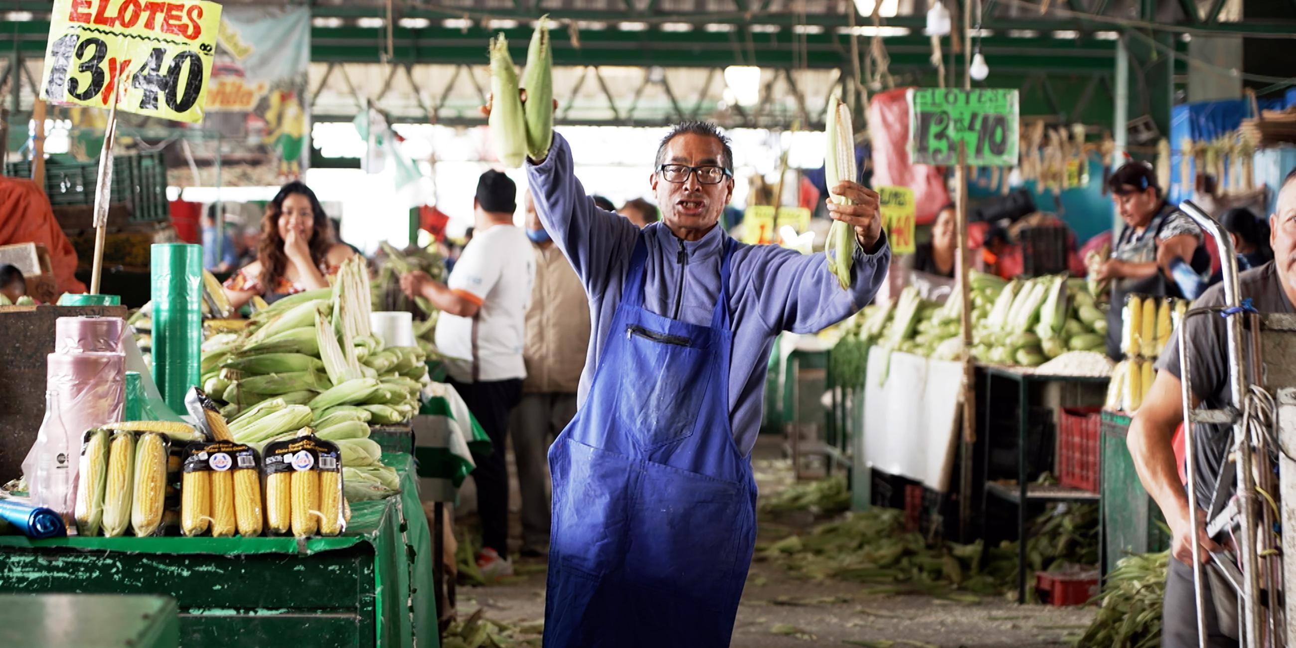 Auf einem Markt: Ein Mannes mittleren Alters trägt einen hellblauen Pullover und eine blaue Schürze. Mit beiden Händen hält er frischen Mais hoch. Um ihn herum stehen unzählige Obst- und Gemüsestände an denen einige Personen stehen.