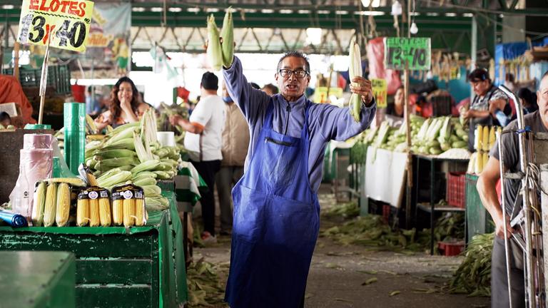 Auf einem Markt: Ein Mannes mittleren Alters trägt einen hellblauen Pullover und eine blaue Schürze. Mit beiden Händen hält er frischen Mais hoch. Um ihn herum stehen unzählige Obst- und Gemüsestände an denen einige Personen stehen.