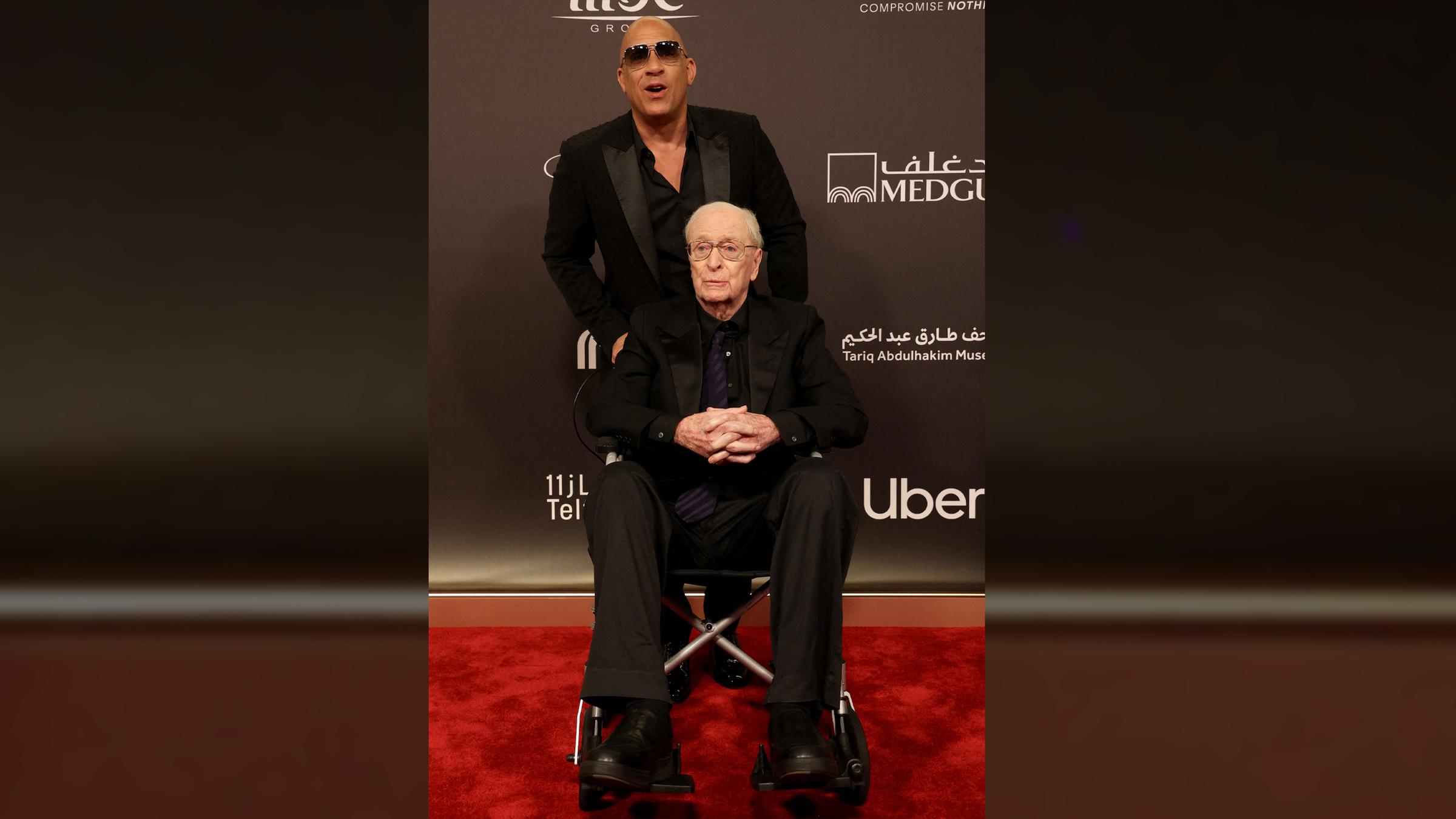 Der Schauspieler Michael Caine sitzt in einem Rollstuhl. Hinter ihm steht der Schauspieler Vin Diesel. Beide tragen schwarze Anzüge, Diesel trägt zusätzlich Sonnenbrille.