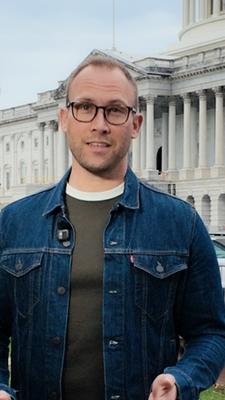 US-Korrespondent David Sauer steht in einer blauen Jeansjacke mit langen Ärmeln auf dem Rasen vor dem US-Kongressgebäude in Washington DC und spricht in die Kamera. Er trägt eine braune Brille.