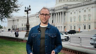 US-Korrespondent David Sauer steht in einer blauen Jeansjacke mit langen Ärmeln auf dem Rasen vor dem US-Kongressgebäude in Washington DC und spricht in die Kamera. Er trägt eine braune Brille.