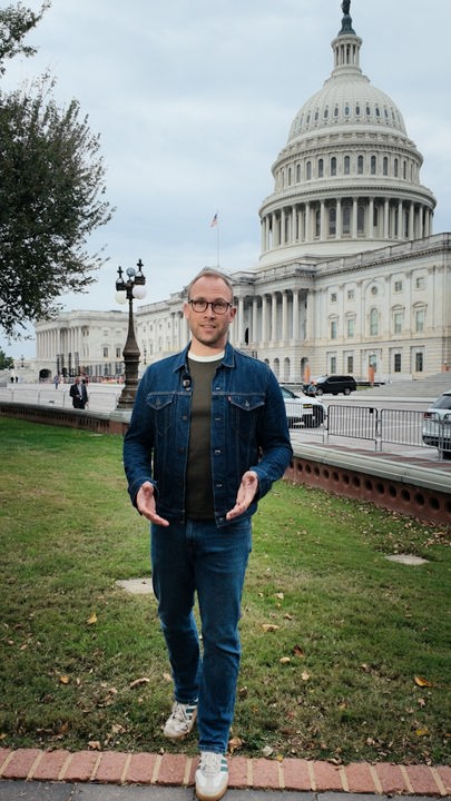 US-Korrespondent David Sauer steht in einer blauen Jeansjacke mit langen Ärmeln auf dem Rasen vor dem US-Kongressgebäude in Washington DC und spricht in die Kamera. Er trägt eine braune Brille.