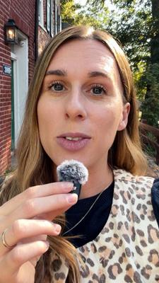 US-Korrespondentin Katharina Schuster steht mit Leoparden-Weste vor einer Hausfassade in Virginia und spricht in ein Mikrofon. Ihre lange braunen Haare fallen ihr über die rechte Schulter. Sie trägt ein dunkles T-Shirt mit weißen Rüschen.