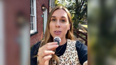 US-Korrespondentin Katharina Schuster steht mit Leoparden-Weste vor einer Hausfassade in Virginia und spricht in ein Mikrofon. Ihre lange braunen Haare fallen ihr über die rechte Schulter. Sie trägt ein dunkles T-Shirt mit weißen Rüschen.