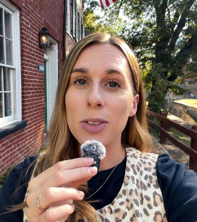 US-Korrespondentin Katharina Schuster steht mit Leoparden-Weste vor einer Hausfassade in Virginia und spricht in ein Mikrofon. Ihre lange braunen Haare fallen ihr über die rechte Schulter. Sie trägt ein dunkles T-Shirt mit weißen Rüschen.