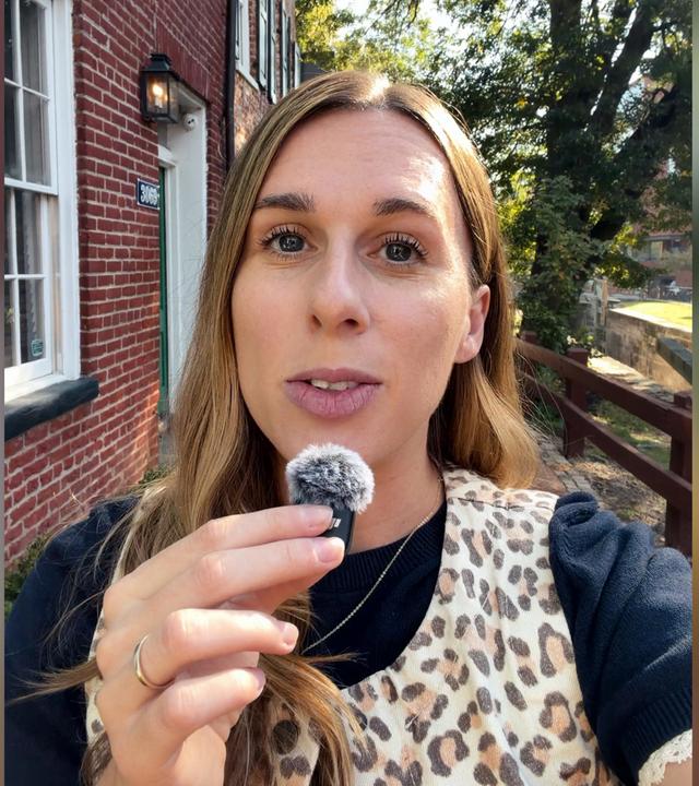 US-Korrespondentin Katharina Schuster steht mit Leoparden-Weste vor einer Hausfassade in Virginia und spricht in ein Mikrofon. Ihre lange braunen Haare fallen ihr über die rechte Schulter. Sie trägt ein dunkles T-Shirt mit weißen Rüschen.