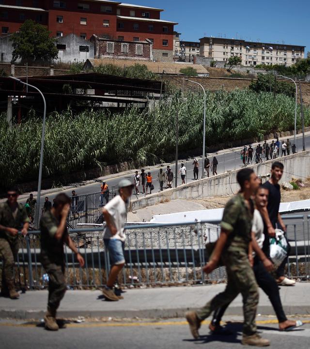 Eine Gruppe von Migranten macht sich am 02.08.2026 in der spanischen Exklave Ceuta in Nordafrika auf den Rückweg von der Kurzzeitaufnahmeeinrichtung CETI in Richtung Grenzübergang nach Marokko, begleitet von spanischen Infanteriesoldaten. 
