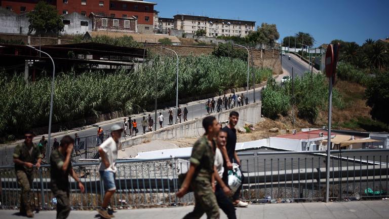 Eine Gruppe von Migranten macht sich am 02.08.2026 in der spanischen Exklave Ceuta in Nordafrika auf den Rückweg von der Kurzzeitaufnahmeeinrichtung CETI in Richtung Grenzübergang nach Marokko, begleitet von spanischen Infanteriesoldaten. 