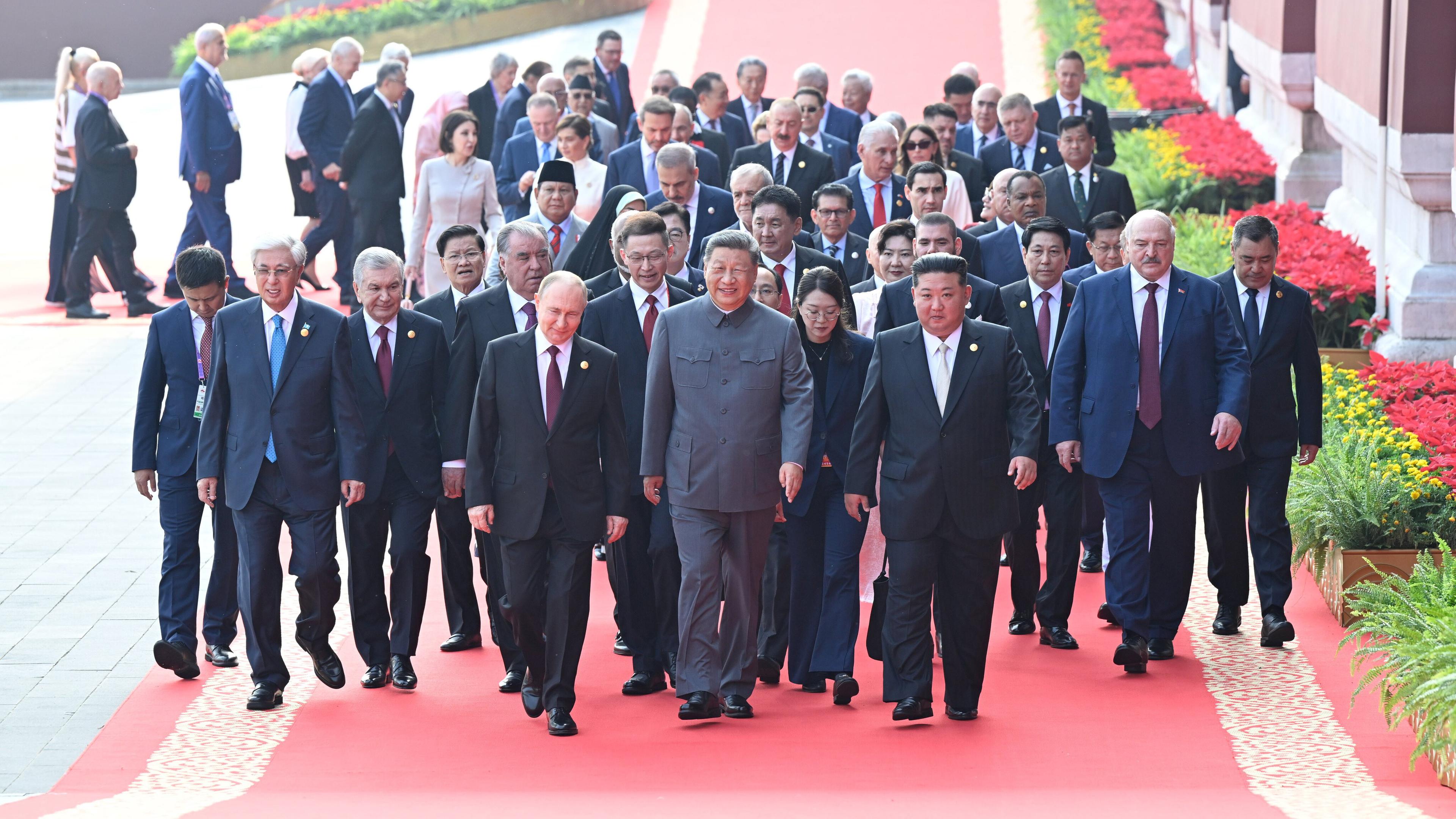 Männer in Anzügen und ein paar Frauen laufen auf einem roten Teppich entlang. In der ersten Reihe laufen Putin (links), Chinas Pärsident Xi (Mitte) und Kim Jong Un (rechts). 