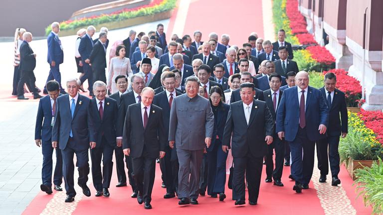 Männer in Anzügen und ein paar Frauen laufen auf einem roten Teppich entlang. In der ersten Reihe laufen Putin (links), Chinas Pärsident Xi (Mitte) und Kim Jong Un (rechts). 