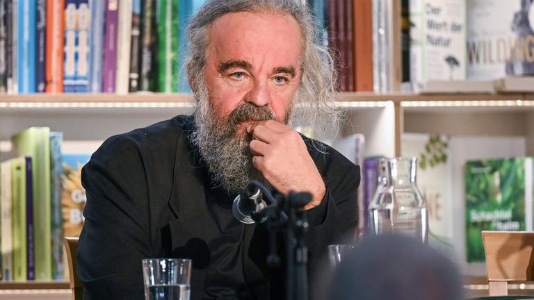 Ein Mann mit graumeliertem Bart und Haaren sitzt an einem Tisch mit Mikrofon und fasst sich nachdenklich mit einer Hand ans Kinn. Hinter ihm befindet sich ein Bücherregal.